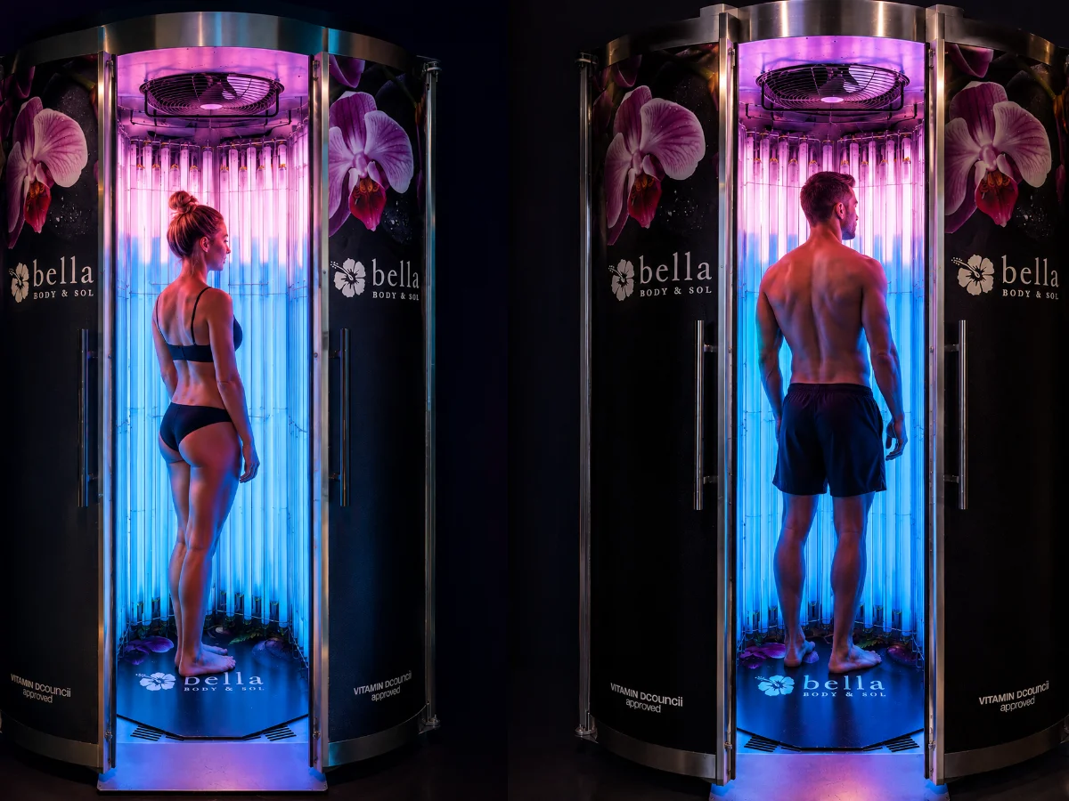 Two people stand inside cylindrical UV tanning booths at a spa, lit by pink and blue lights, with Bella Body & Sol branding visible on the walls.